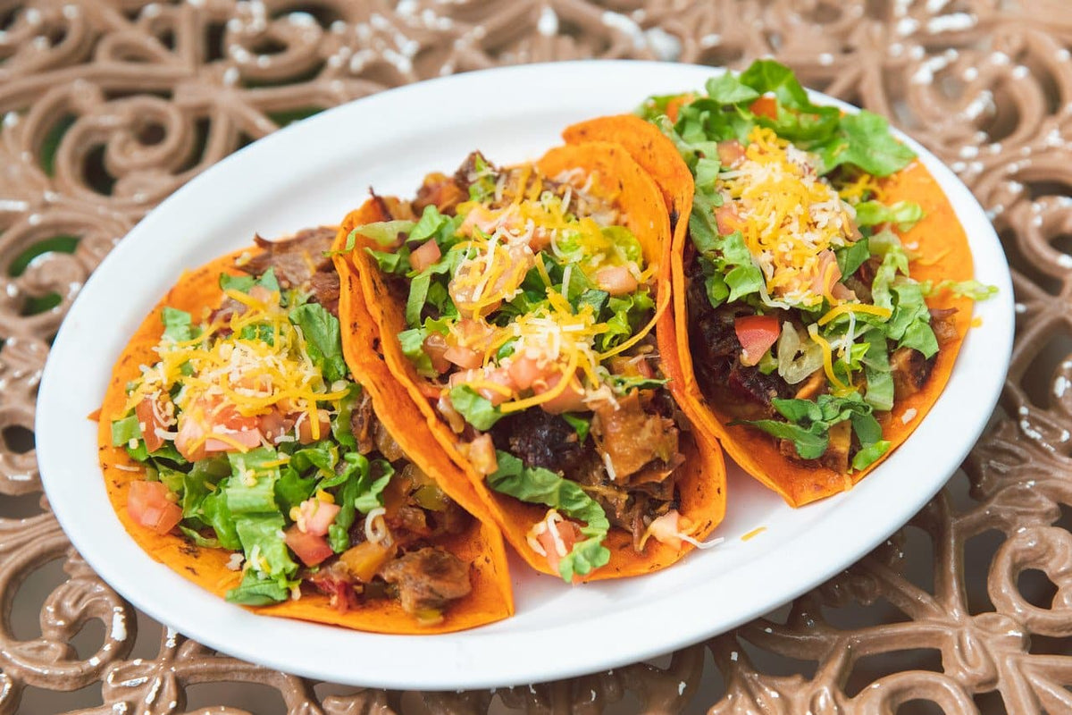 Three tacos made with red chile corn tortillas, filled with seasoned meat, lettuce, tomato and cheese