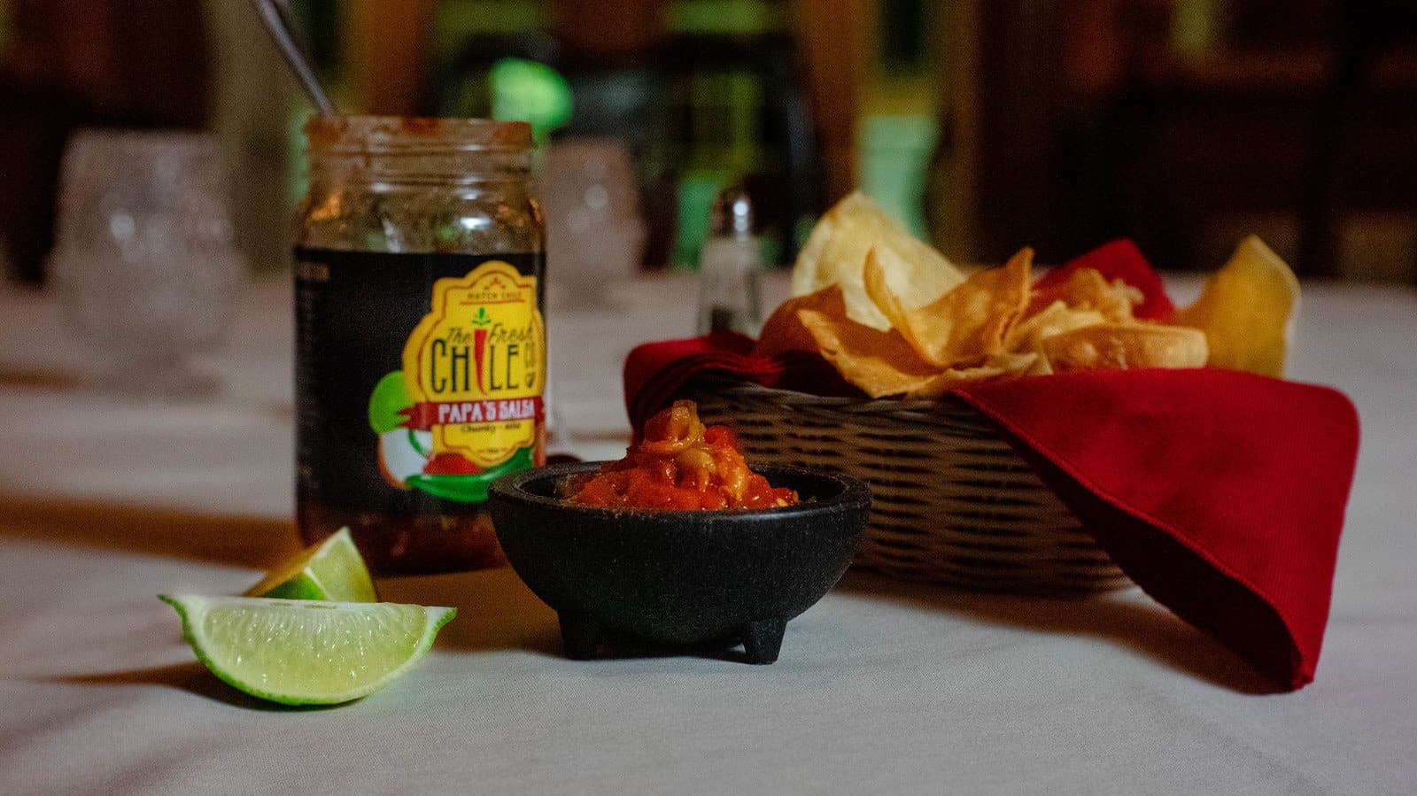 A small bowl of Fresh Chile Co's Mama's Blended Hatch Chile Salsa next to a basket of tortilla chips, with a lime wedge and a jar labeled "chiles" on a dimly lit table