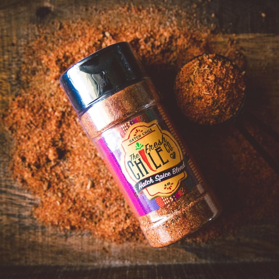 A bottle of "Hatch Red Chile Spice Blend" on a wooden table, surrounded by scattered chili flakes. The label features pink and yellow colors.