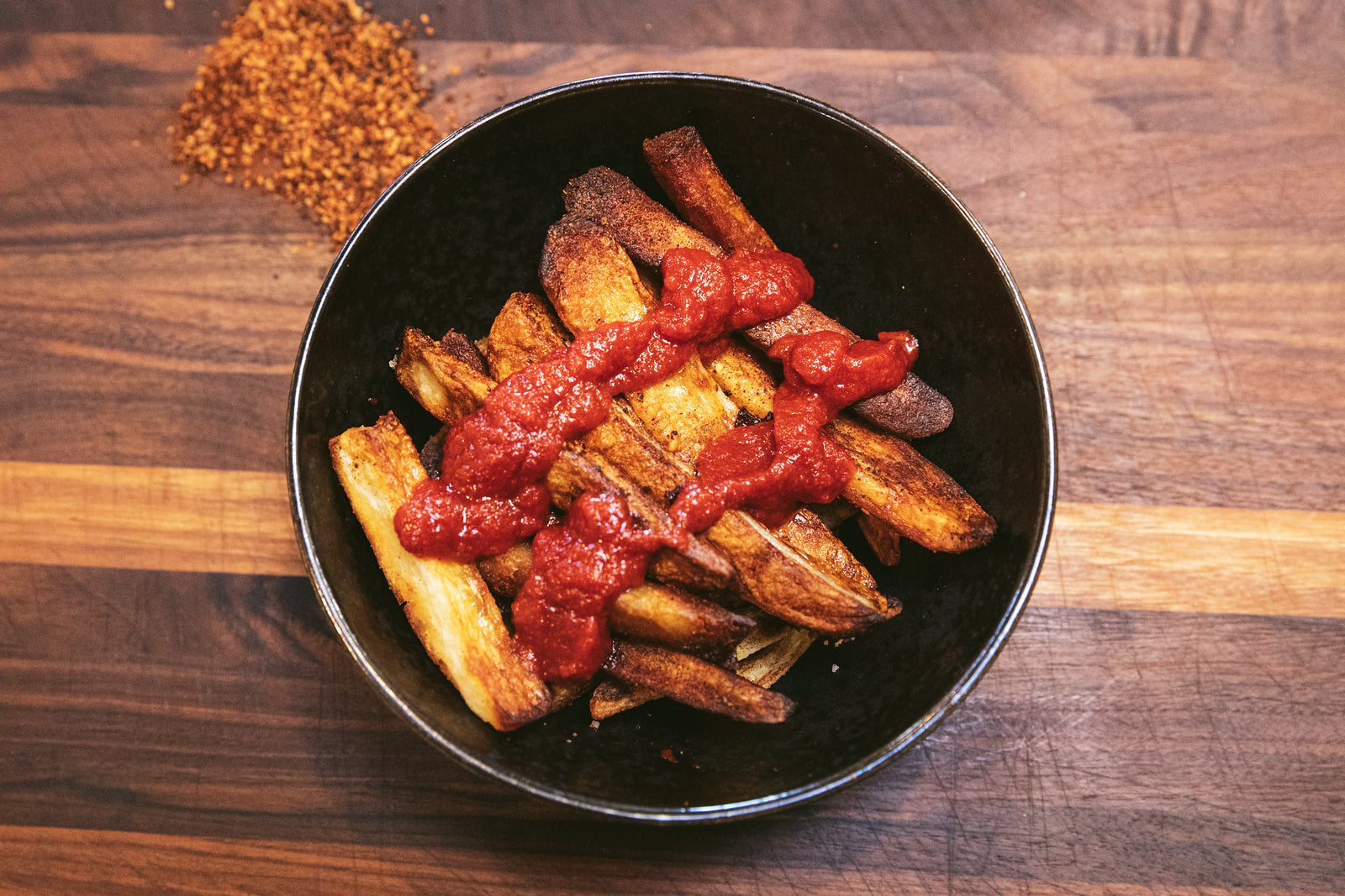 A bowl of crispy potato wedges topped with Hatchup - Spicy Hatch Red Chile Ketchup, sitting on a wooden table, with a sprinkle of crushed red pepper in the corner.