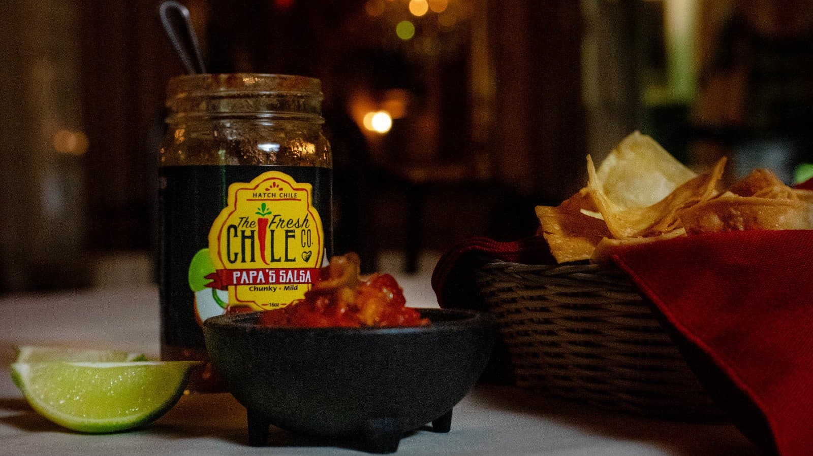 A jar of Papa's Chunky Hatch Chile Salsa beside a bowl of salsa and tortilla chips, with a lime wedge on a table, illuminated in dim, warm.