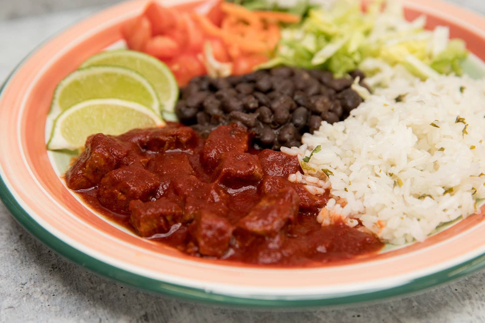 A plate of Mexican food featuring chicken enchiladas in Sun-Dried Red Chile Sauce, accompanied by white rice, black beans, diced tomatoes, shredded lettuce, and slices of lime.