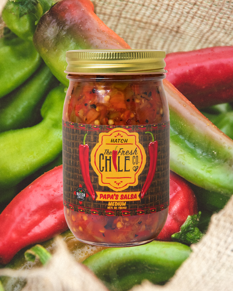 A jar of "Hot Salsa Sampler" medium spiciness, surrounded by fresh green and red chili peppers, placed on a burlap surface. The label features a graphic of a red chili.