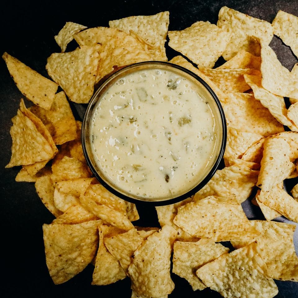 A bowl of creamy white Pure Hatch Green Chile queso dip surrounded by scattered tortilla chips on a dark surface.