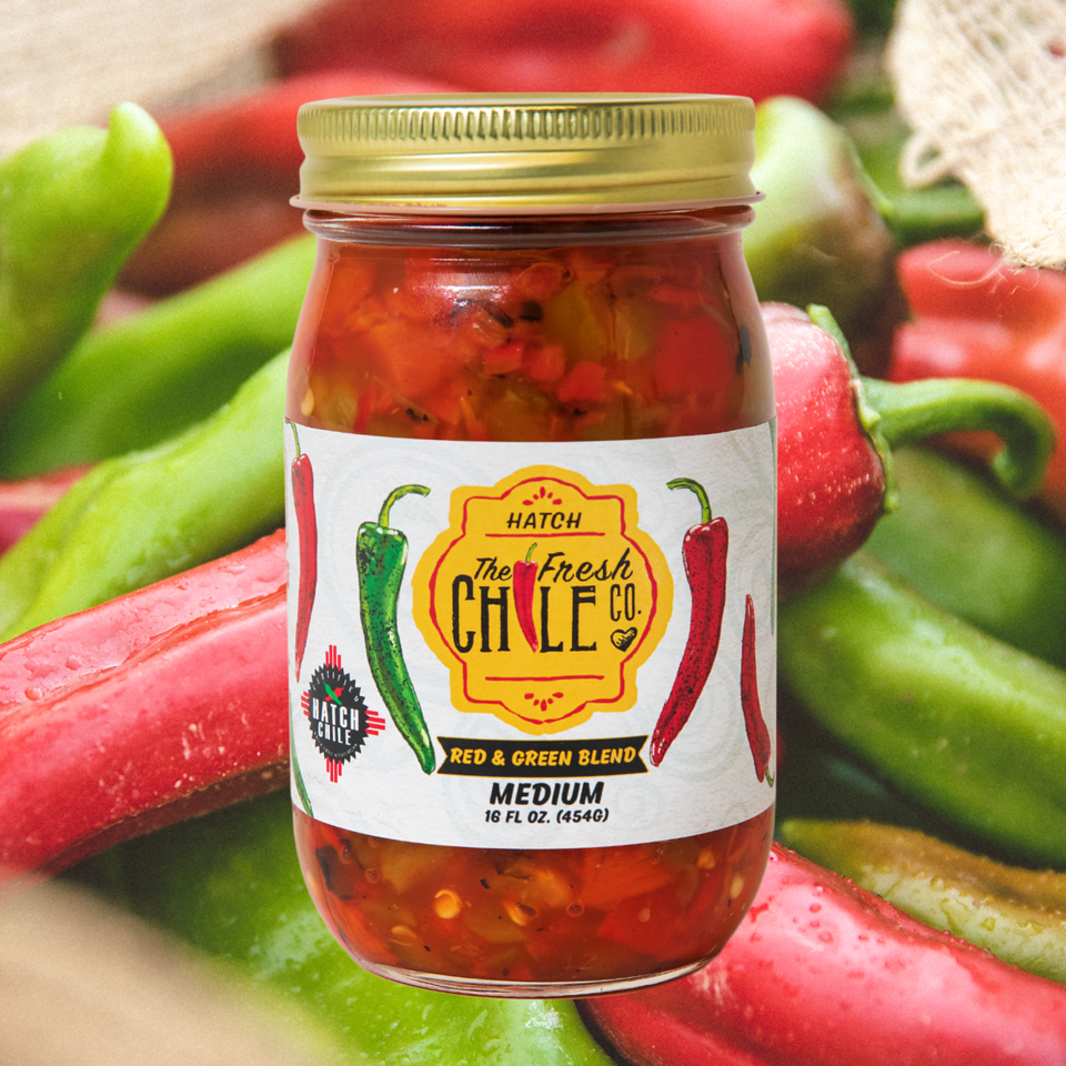 A glass jar of Sauces & Roasts Sampler surrounded by fresh red and green chili peppers on a textured surface, perfect for variety gift packs.