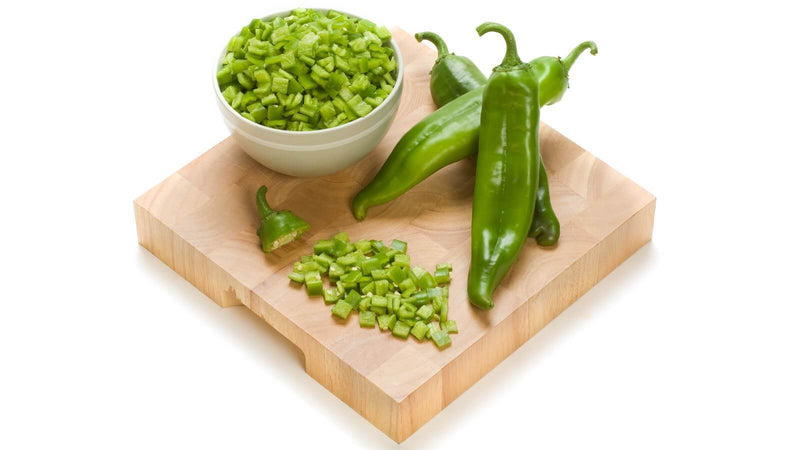 A white bowl of diced green chile and whole green chile pods on a wooden cutting board against a white background