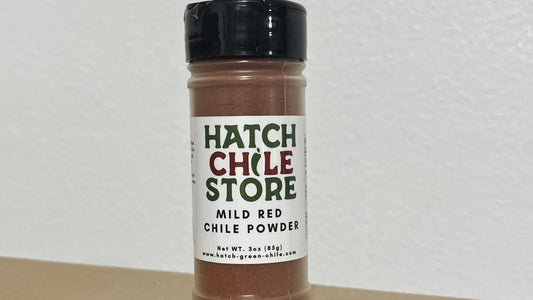 A shaker jar of Hatch Chile Store Mild Red Chile Powder, 3 oz, standing on a table against a white wall