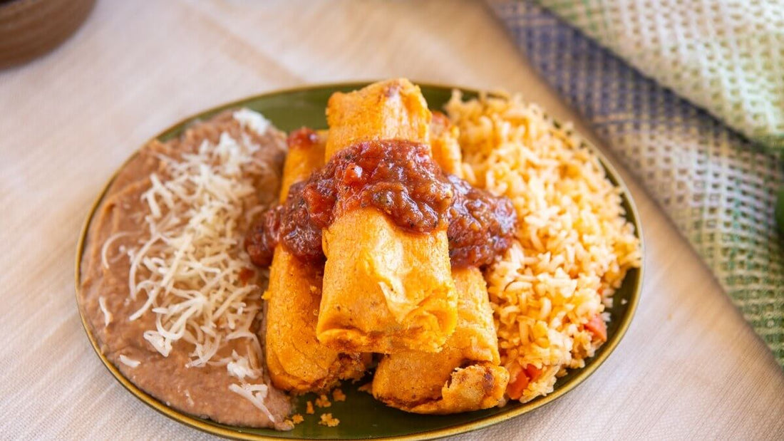 Unwrapped tamales topped with red chile salsa on a green plate, with refried beans and shredded cheese on one side and Spanish rice on the other