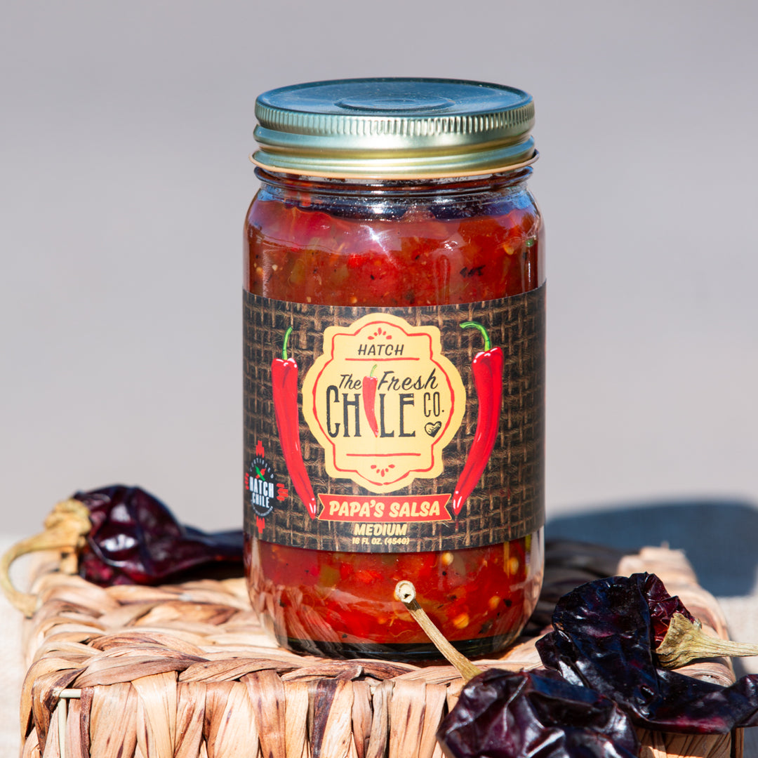 A glass jar of Papa's Chunky Hatch Chile Salsa labeled "papa’s salsa medium," surrounded by dried chilies on a woven basket with a neutral background.