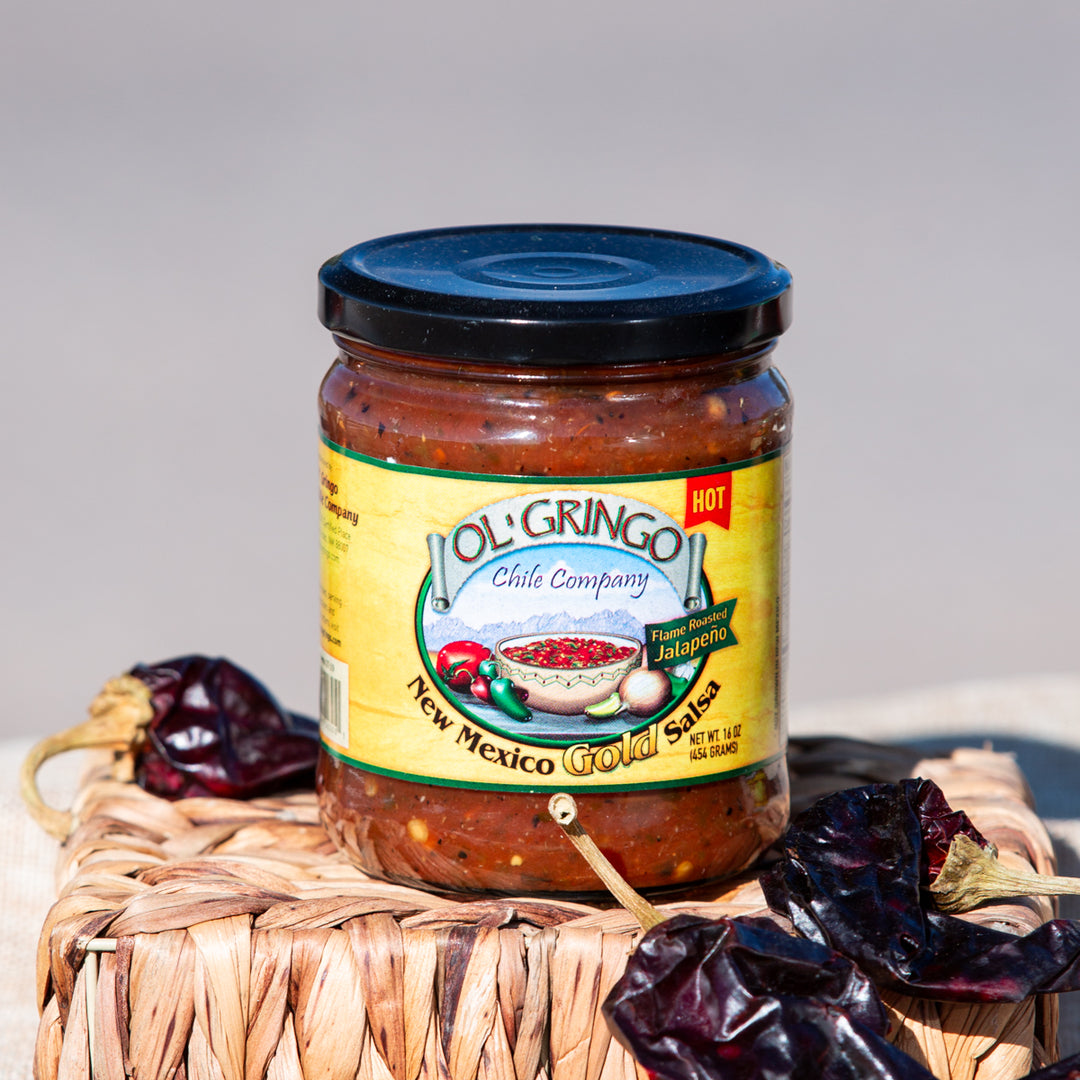 A jar of New Mexico Gold Salsa, with a label featuring red and green chiles, placed on a woven mat next to dried chiles, under bright natural