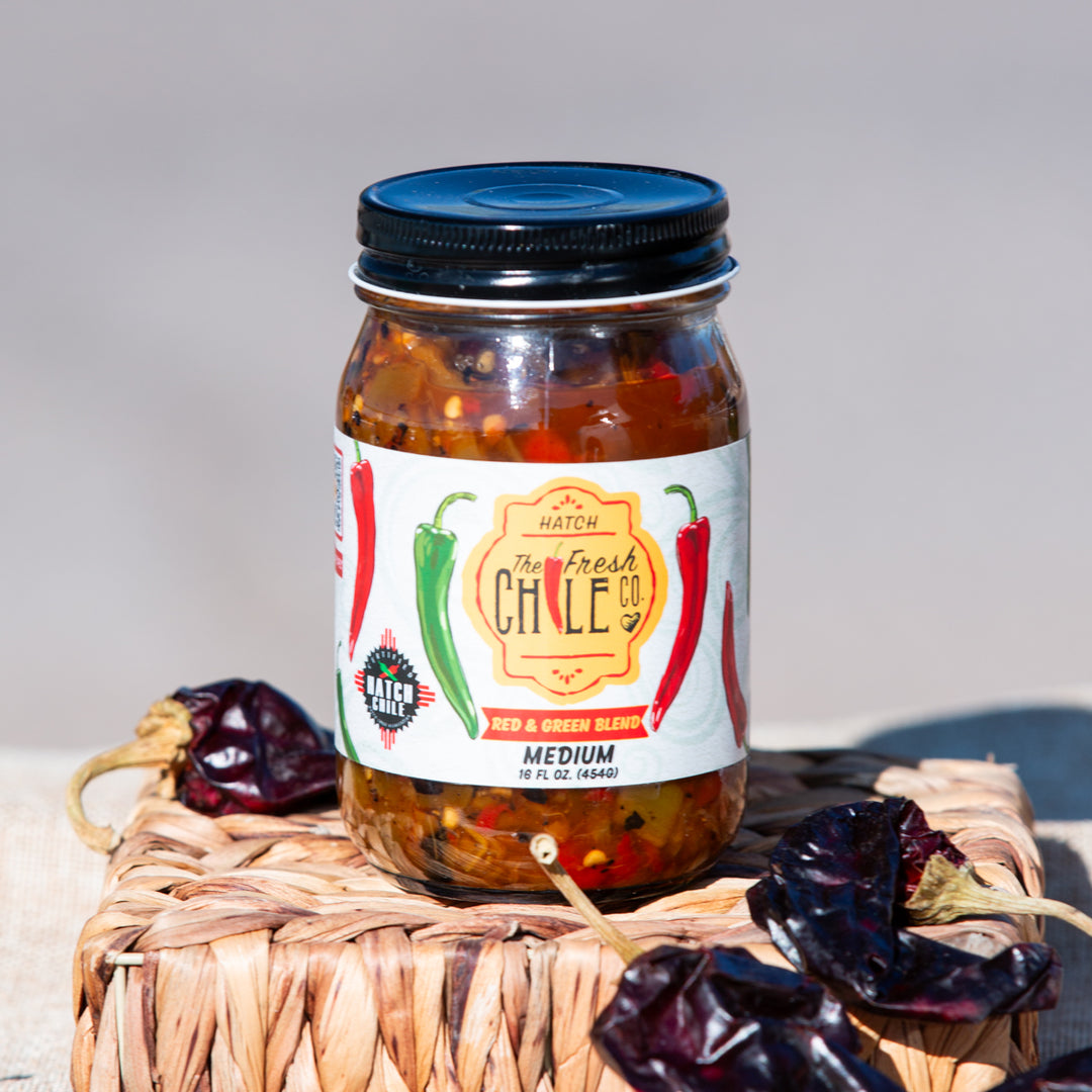 A jar of Hatch red & green chile salsa sits on a woven basket, accompanied by dried chili peppers, against a neutral background.