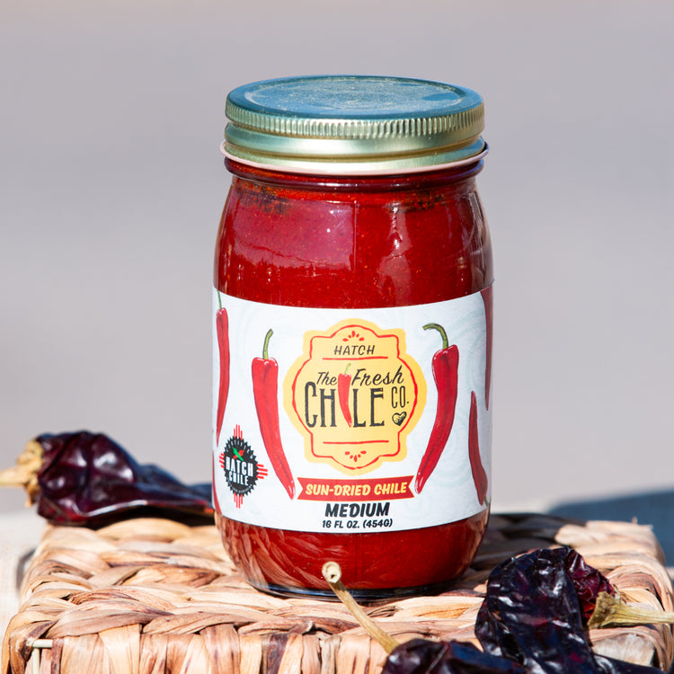 A jar of Sun-Dried Red Chile sauce labeled "medium," placed on a woven mat with dried chilies scattered around, under natural lighting.