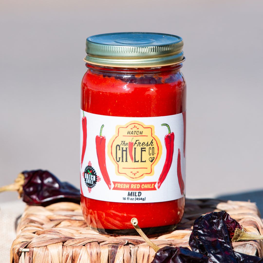 A jar of Fresh Hatch Red Chile Sauce by the Hatch Chile Co., gluten free and flanked by dried chiles, sits on a wicker mat with a clear blue sky in the background.