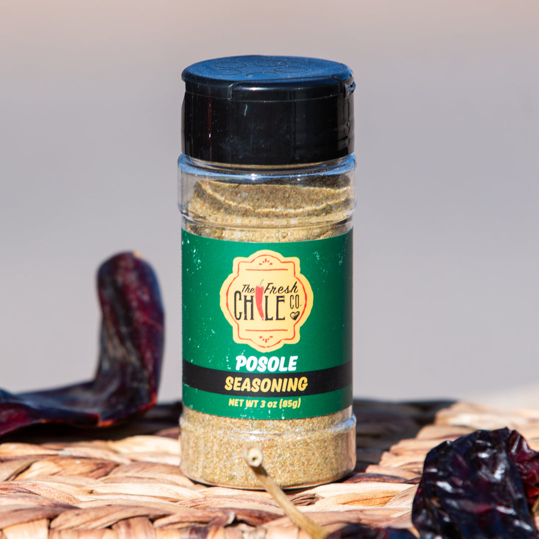 A jar of Posole Pack seasoning labeled "the fresh chile co." on a woven mat, with dried chili peppers and white hominy around it, showcasing the key ingredient under natural light.