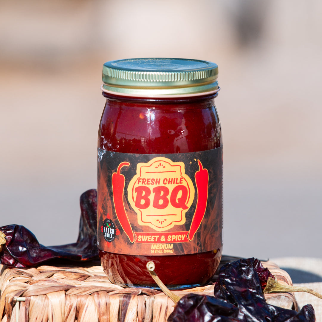 A jar of fresh Sweet & Spicy Hatch Red Chile BBQ sauce sits on a woven basket surrounded by dried chiles. The jar has a red label and a green lid.