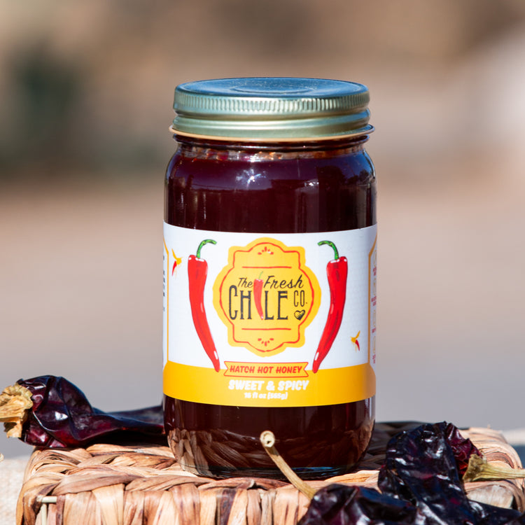 A jar of Hatch Hot Honey, labeled sweet and spicy, sits on a basket surrounded by dried red chilies against a blurred natural background.