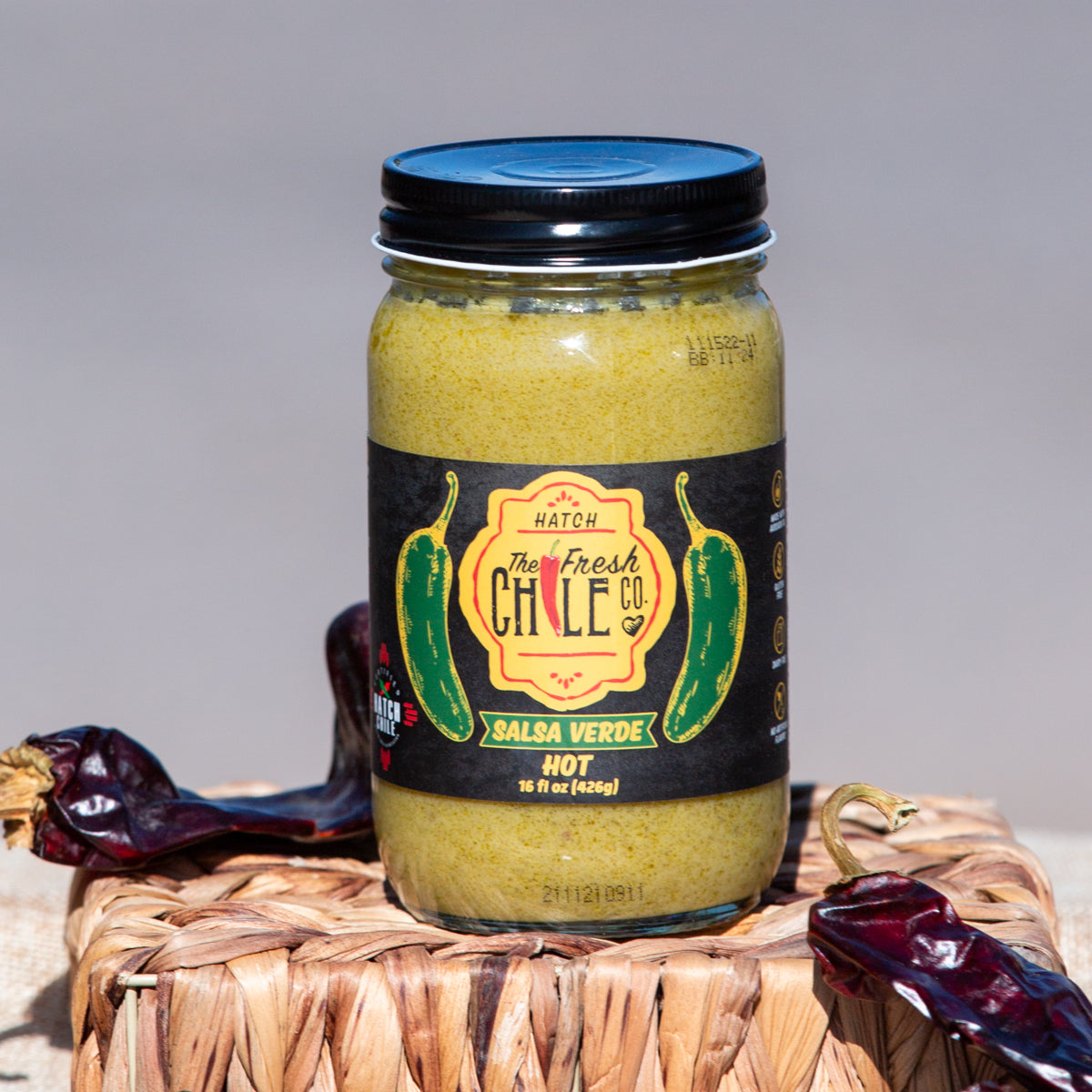 A jar of Hatch Jalapeño Salsa Verde on a wicker mat, flanked by dried red chiles, with a neutral beige backdrop.