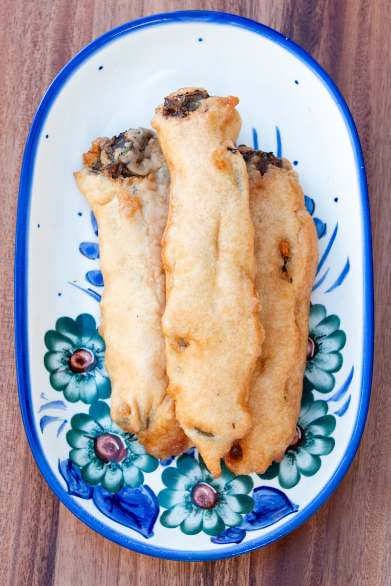 Three golden-brown fried spring rolls filled with vegetables and Hatch Chile Rellenos on a floral oval plate, viewed from above on a wooden table.