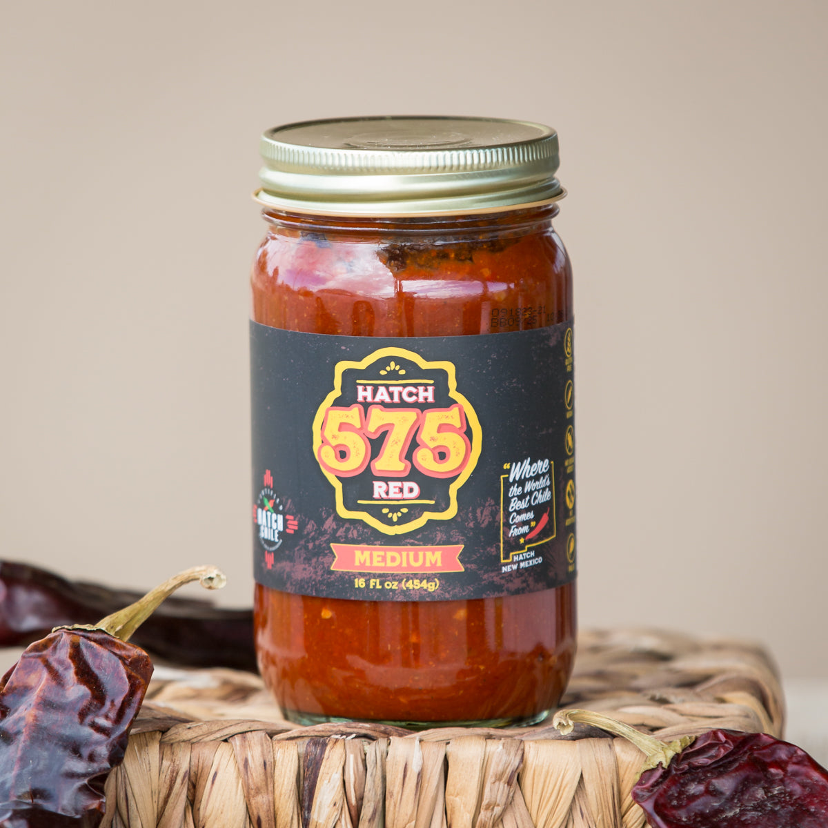 A jar of Hatch 575 Red Chile Sauce, labeled "medium" heat, placed on a woven basket with scattered dry chili peppers around it. The jar is sealed with a metal lid.