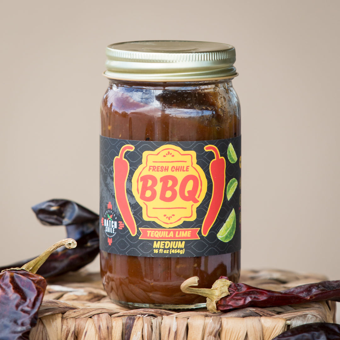 A jar of Hatch Tequila Lime BBQ sauce with a vibrant label, surrounded by dried Hatch chiles on a woven basket, set against a neutral background.