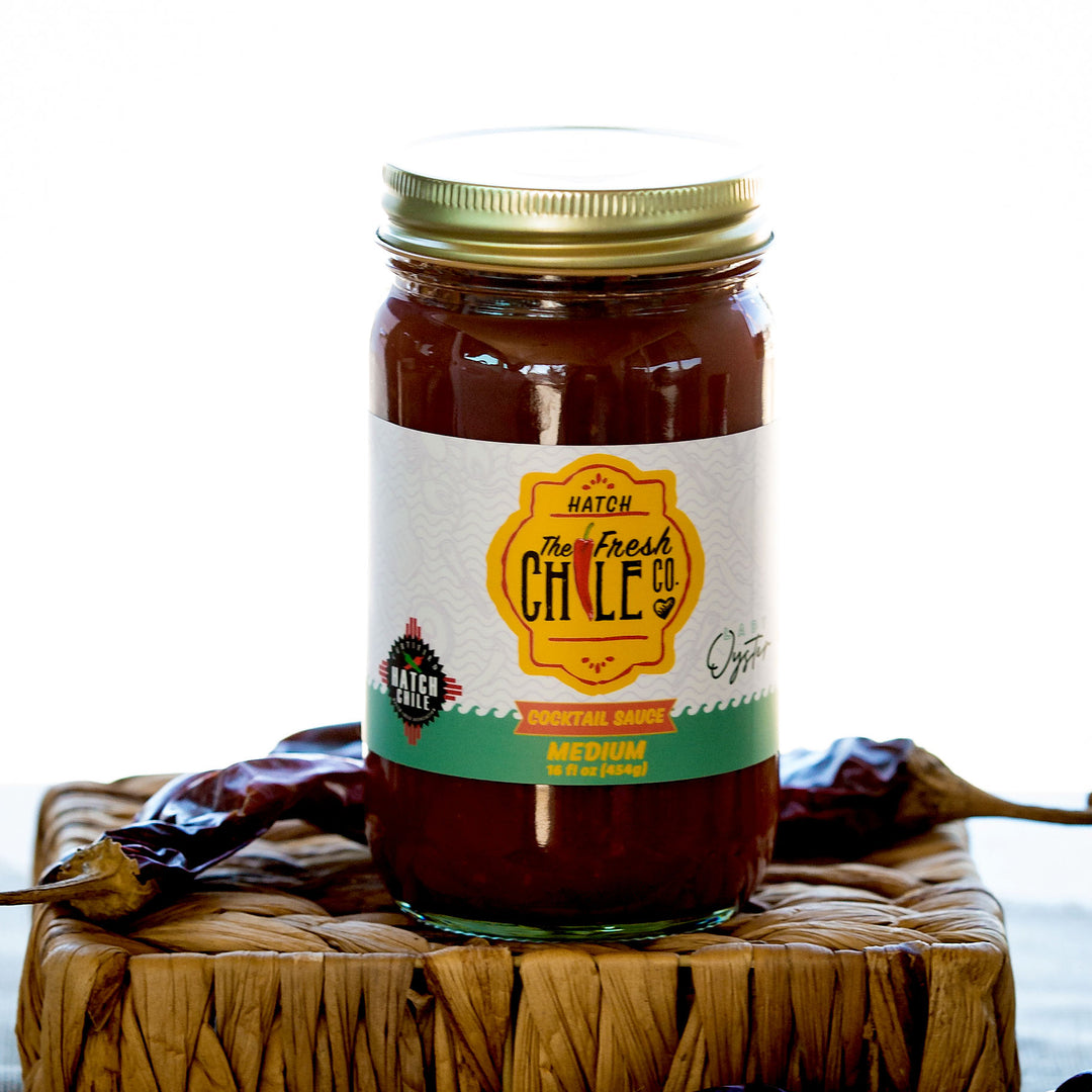 A jar of Hatch Chile Cocktail Sauce on a woven mat, flanked by dried chilies, with a blurred background emphasizing the product.