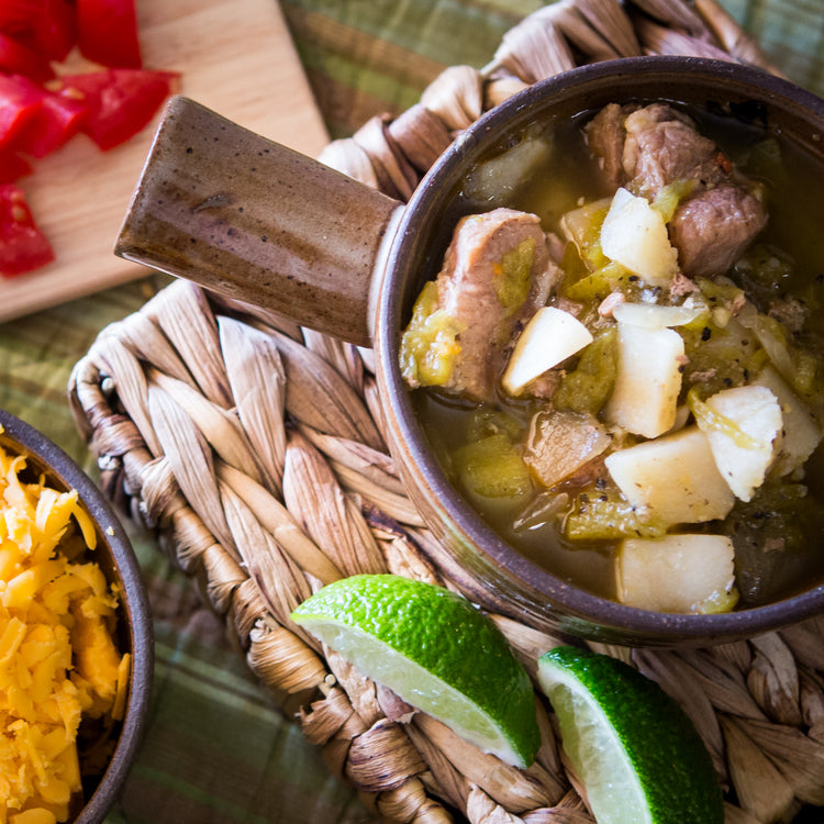 A bowl of traditional **Hatch Green Chile Stew** with chunks of pork and potatoes, garnished with diced onions, accompanied by lime wedges and a side of shredded cheese on a woven placemat.