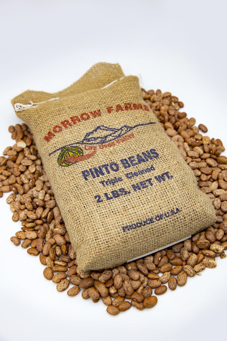 A burlap sack labeled "Morrow Farms Pinto Beans, 2 lbs. net wt., produce of USA" rests on a pile of pinto beans against