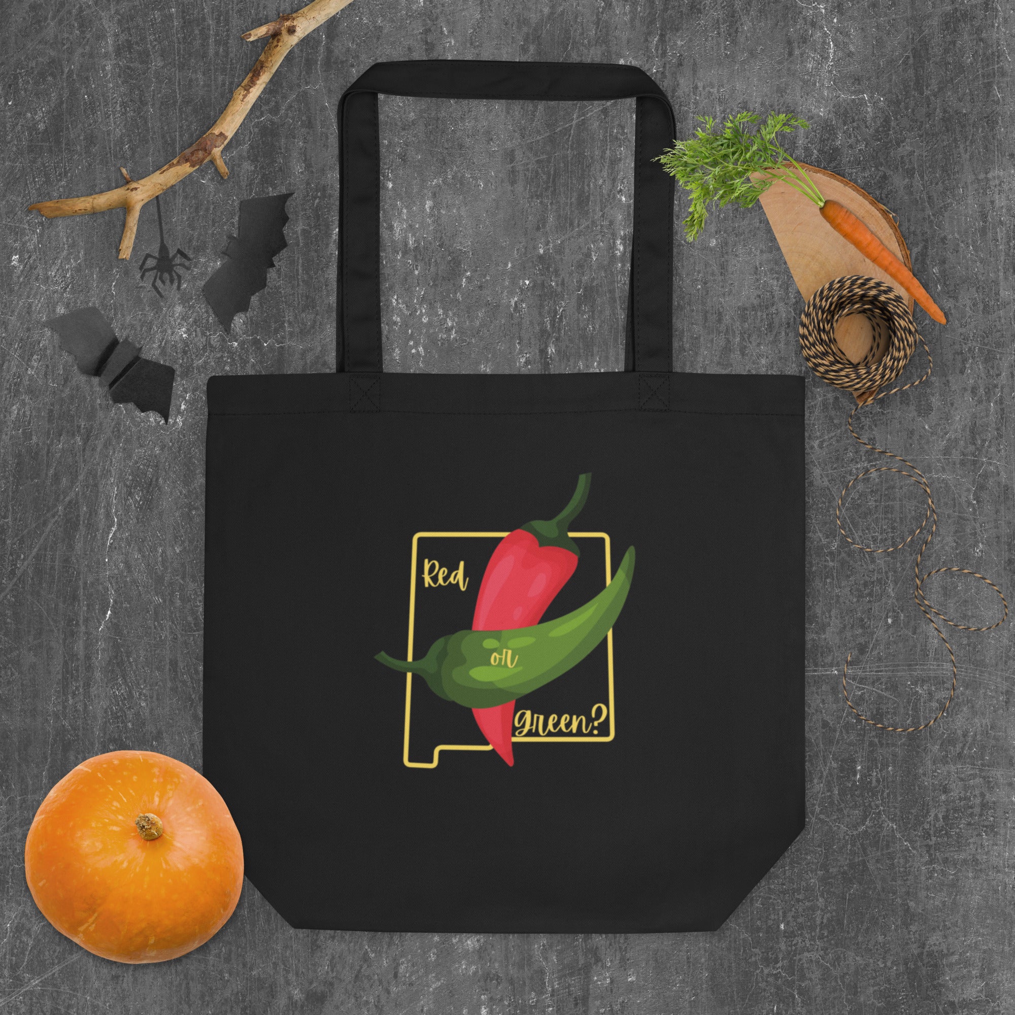 A NM State Question Tote Bag with a design featuring a red and a green chili pepper on a yellow background, captioned "red or green?". The bag is displayed on a gray surface, surrounded