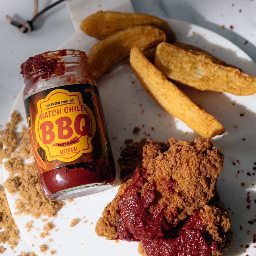 A jar of "Sweet & Spicy Hatch Red Chile BBQ Sauce" in medium heat beside crispy fried chicken and potato wedges, on a white plate with scattered sauce and crumbs.