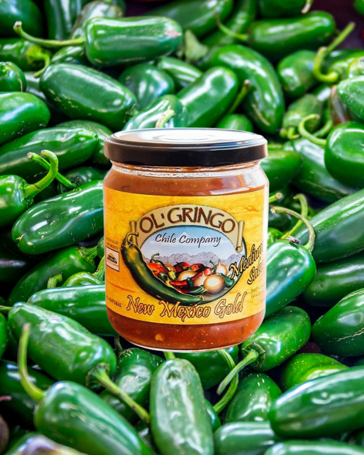 A jar of Hot Salsa Sampler labeled "new mexico gold" surrounded by fresh green jalapeño peppers. The jar stands out with its bright orange label and is part of a