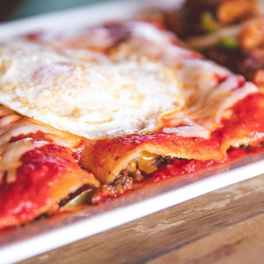Close-up view of a gluten-free lasagna dish topped with a fried egg, showing layers of pasta, meat, and Fresh Hatch Red Chile Sauce on a white plate.