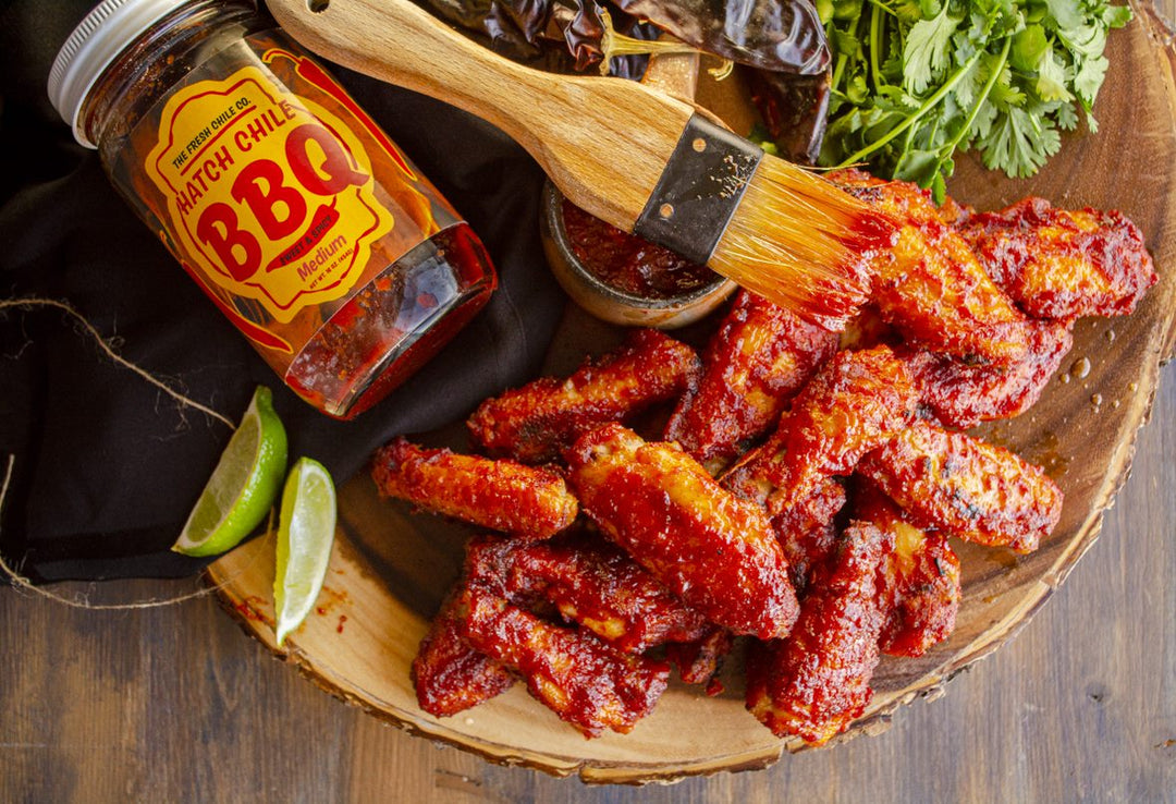 A wooden bowl filled with spicy barbecue chicken wings, accompanied by a bottle of Sweet & Spicy Hatch Red Chile BBQ Sauce, a brush, lime wedges, and fresh cilantro on a rustic table.