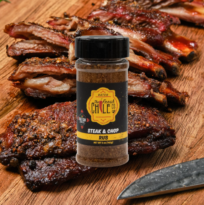 A bottle of Hatch Red Chile Steak & Chop Rub stands next to sliced grilled meat and a knife on a wooden cutting board.