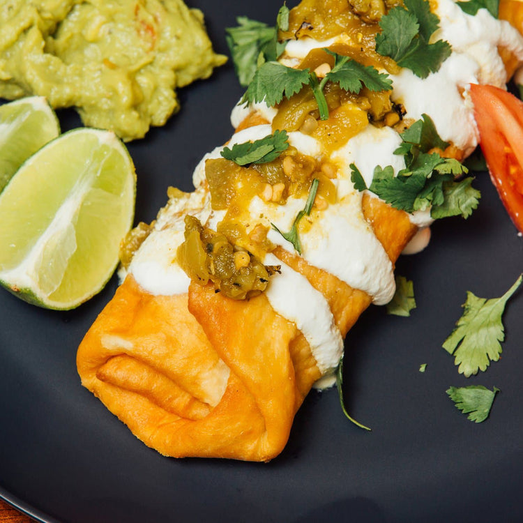 A plate with a Hatch Green Chile Beef & Cheese Chimichanga topped with white sauce, pickled jalapeños, mango salsa, and cilantro alongside guacamole and lime wedges.