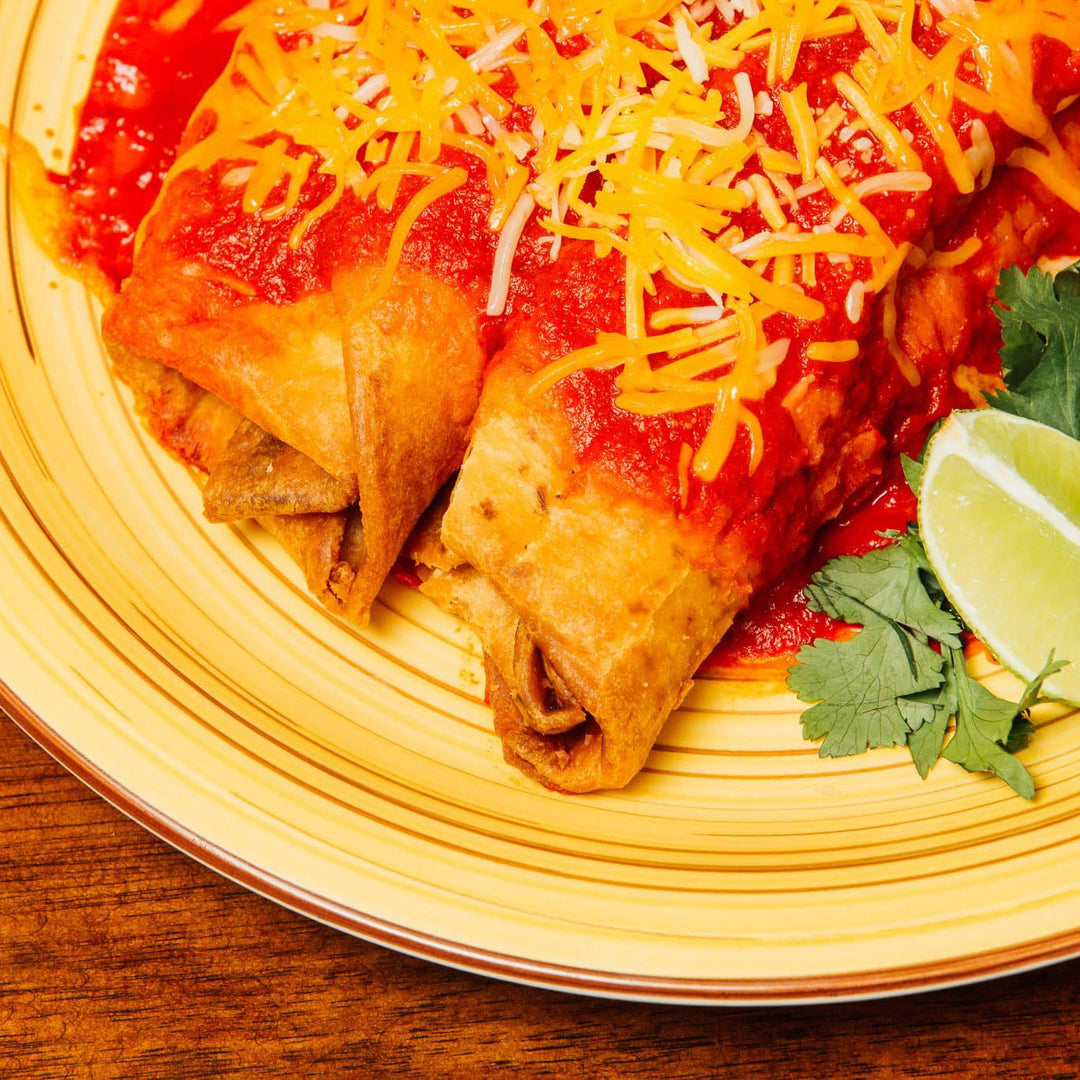 Close-up of a plate with two Hatch Green Chile Beef & Cheese Chimichangas topped with Hatch Green Chile sauce and shredded cheese, garnished with a lime wedge and fresh cilantro, on a wooden table.