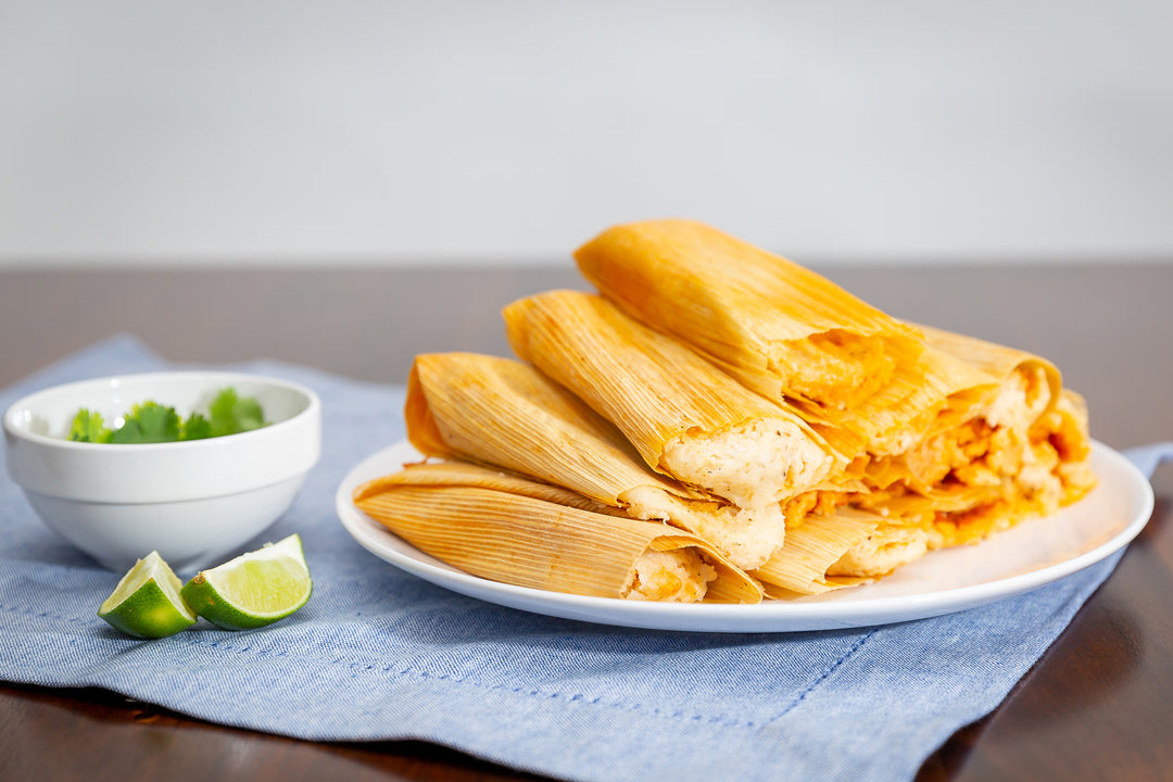 A plate of freshly made Meat Lover Sampler, served with lime wedges and salsa on a wooden table. The tamales are neatly placed on a white plate over a blue napkin.