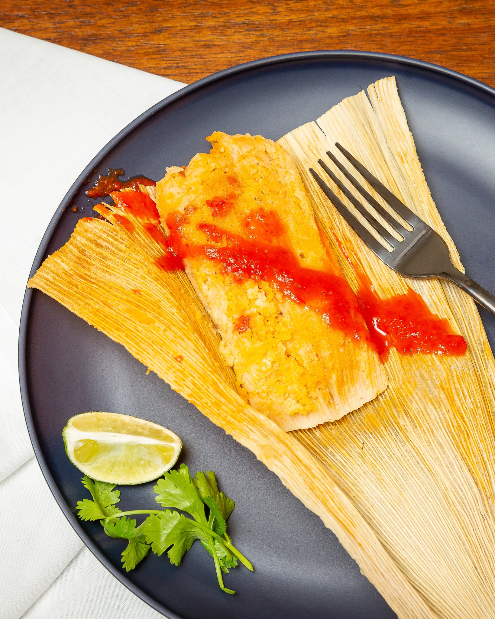 A Meat Lover Sampler served on a corn husk, drizzled with red sauce, on a dark blue plate with a slice of lime and cilantro on the side, and a fork resting on the plate.