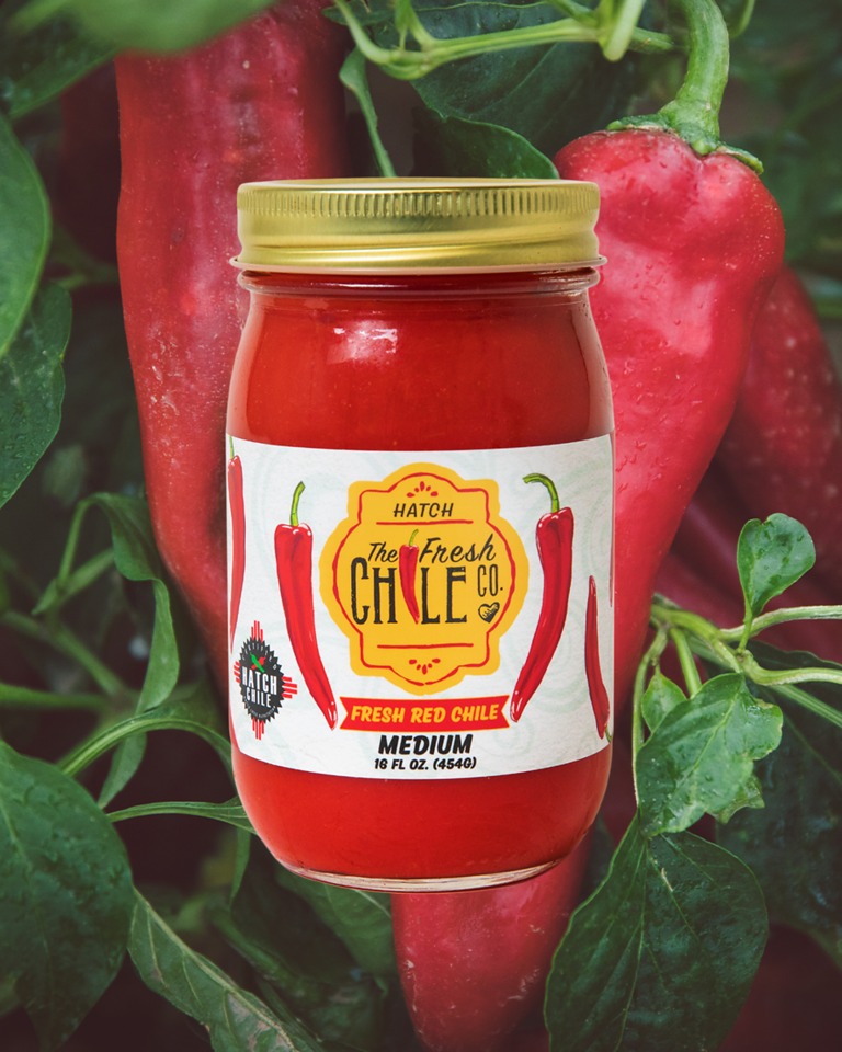 A jar of Sauces & Roasts Sampler labeled "medium" surrounded by fresh red chilies and green foliage, emphasizing the product's natural and spicy ingredients.