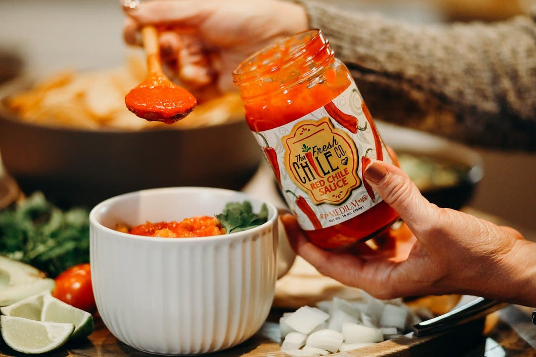 A person scooping Fresh Hatch Red Chile Sauce from a jar labeled "farm fresh Fresh Hatch Red Chile Sauce" into a white bowl on a table with fresh vegetables and tortilla chips.