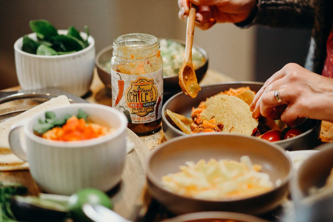 Hands preparing tacos with various ingredients on a table, including a jar of Hatch Red & Green Chile sauce, fresh greens, sliced tomatoes, and shredded cheese.