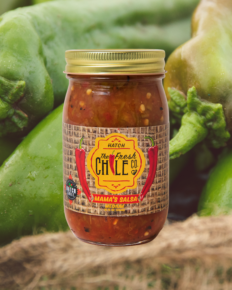 A jar of Hot Salsa Sampler labeled "the fresh chile co." sits in front of several fresh green chile peppers, suggesting the ingredients of the salsa.