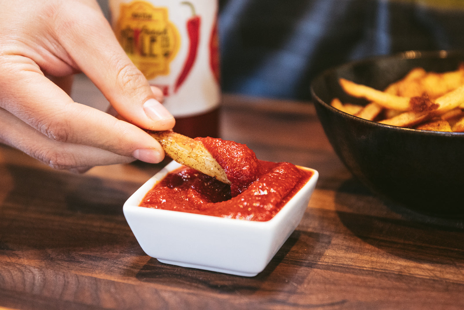 A person dips a seasoned french fry into a small white bowl of Hatchup, with a soda can and another bowl of fries in the background on a wooden table.