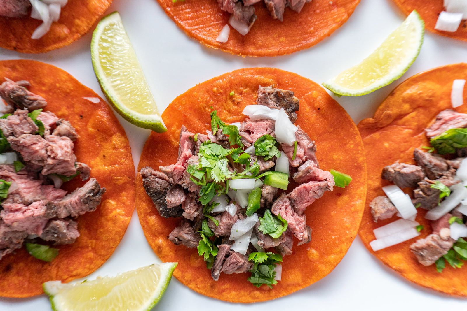 Grilled steak tacos on Hatch Pueblo Bread Red Chile Corn Tortillas, garnished with chopped onions, cilantro, and lime wedges on a white plate.